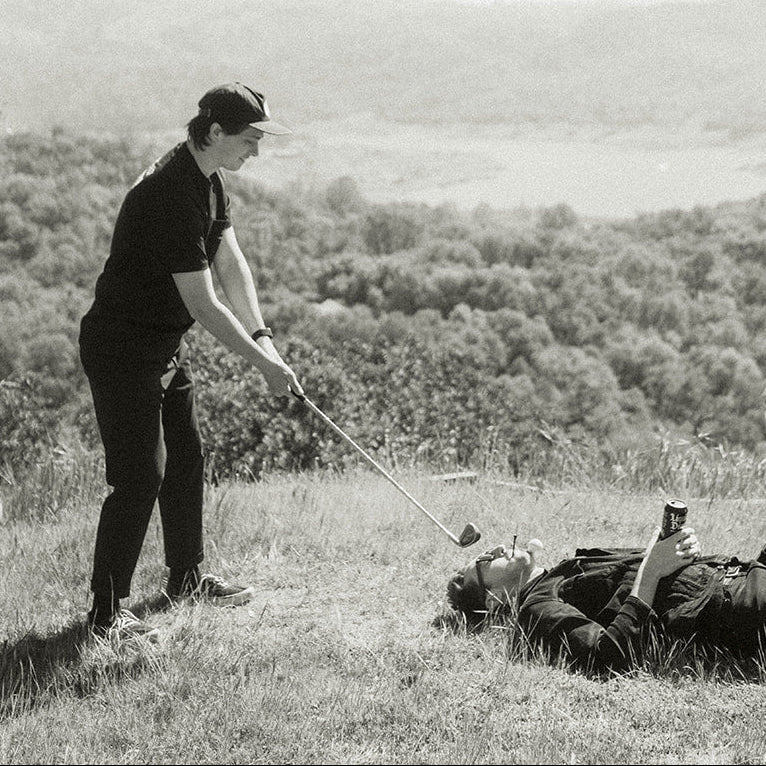 Person playing golf with another person lying on the ground in a scenic outdoor setting.